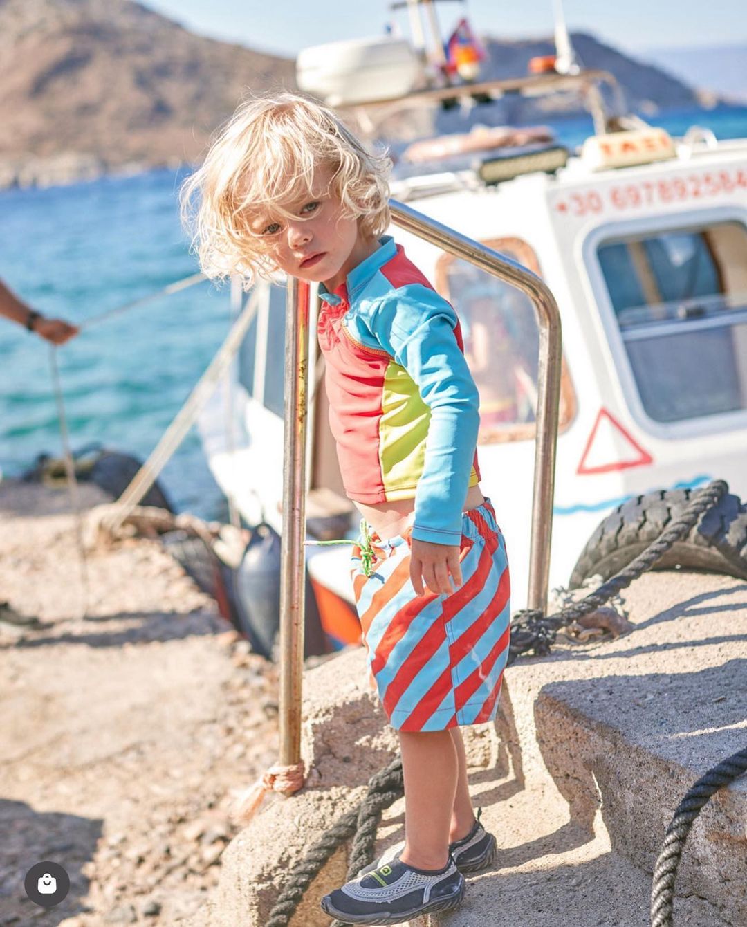 Young boy with blond hair stands on a dock wearing a colorful rash guard and striped shorts in front of a boat and ocean.