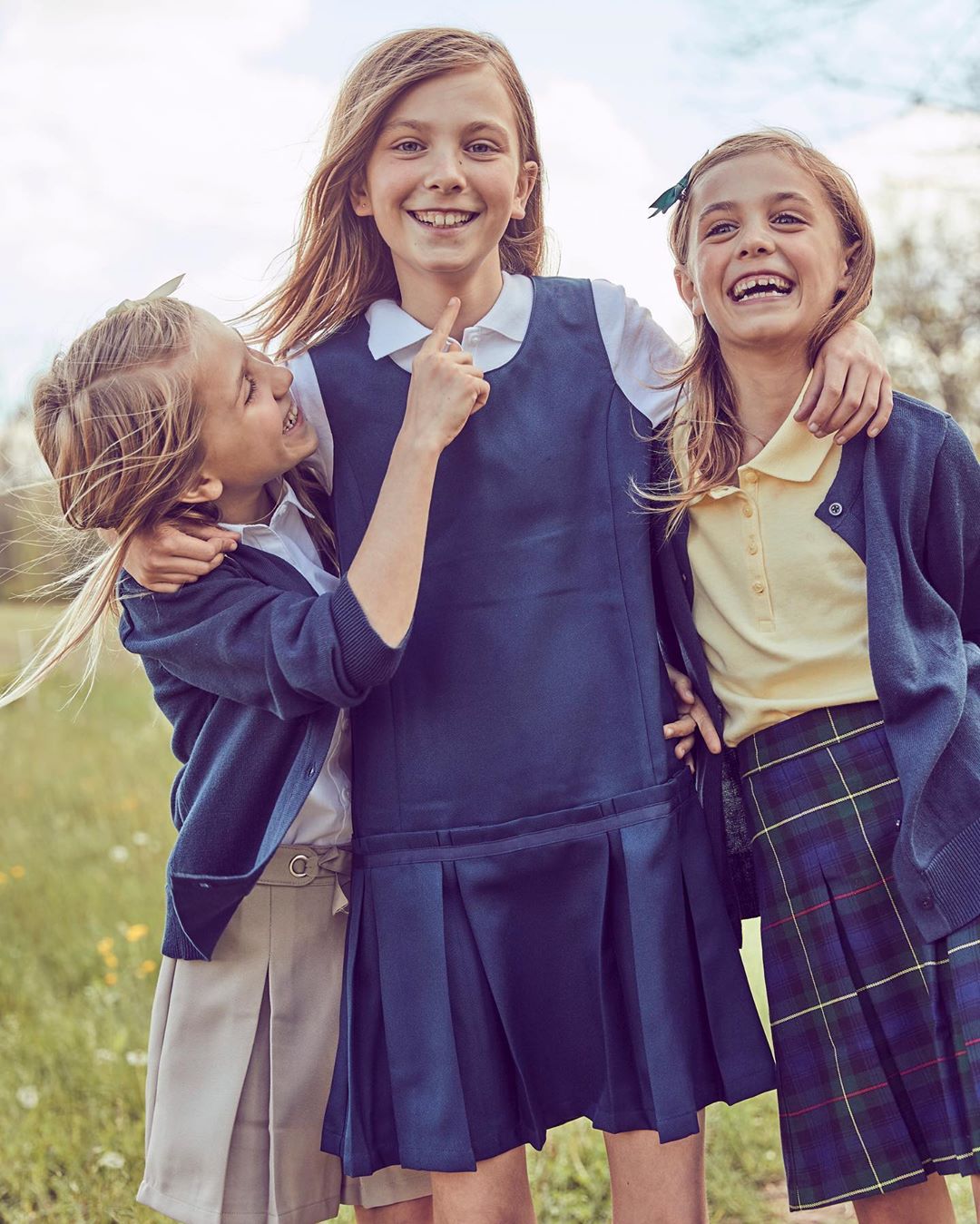 Three smiling young girls in school uniforms embrace outdoors. One girl points upward, while others wear cardigans and pleated skirts.