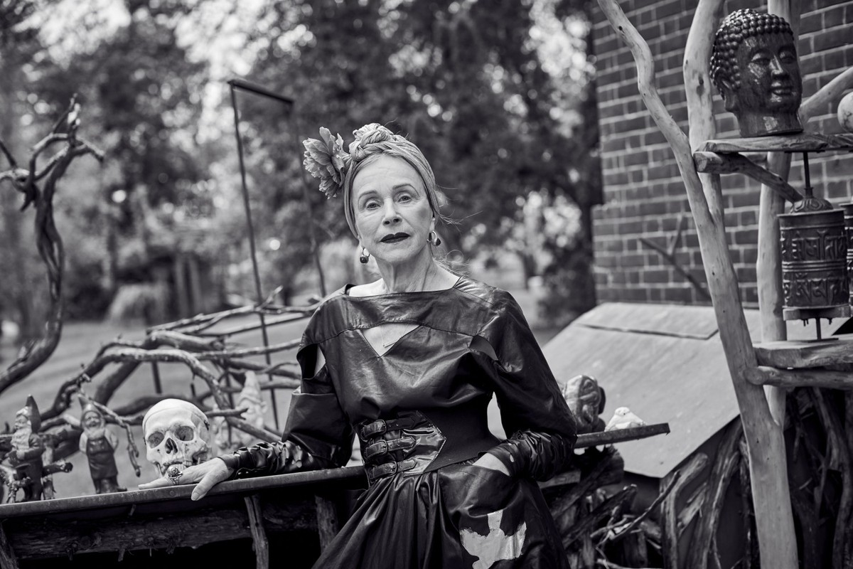 Black-and-white portrait of a senior woman with a head wrap, wearing a dark dress, posed with a skull and garden gnomes outdoors.