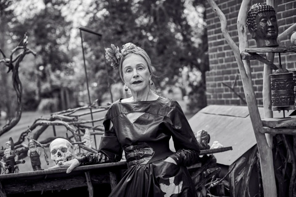 Black-and-white portrait of a senior woman with a head wrap, wearing a dark dress, posed with a skull and garden gnomes outdoors.