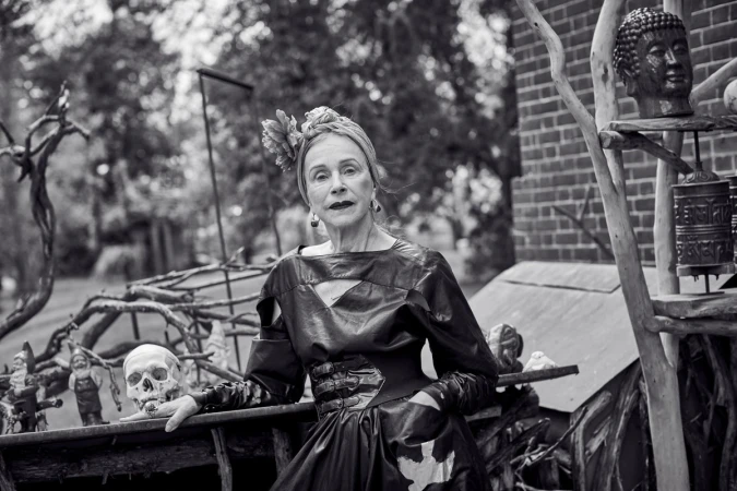 Black-and-white portrait of a senior woman with a head wrap, wearing a dark dress, posed with a skull and garden gnomes outdoors.