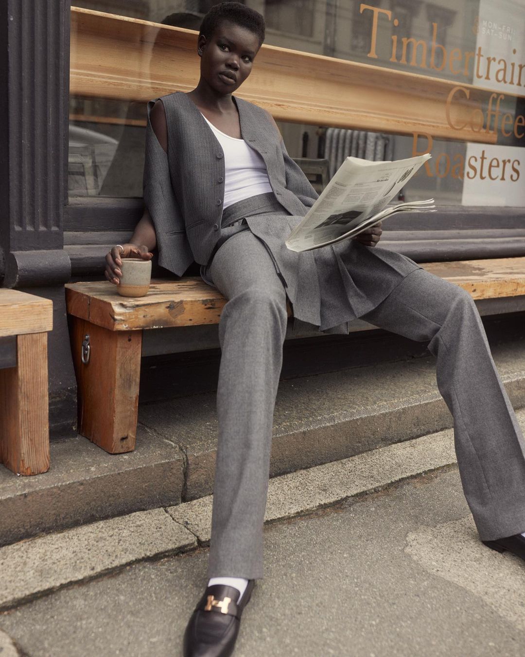 A woman in a grey suit sits on a bench outside, reading a newspaper and drinking coffee. Cafe sign is visible in the background.