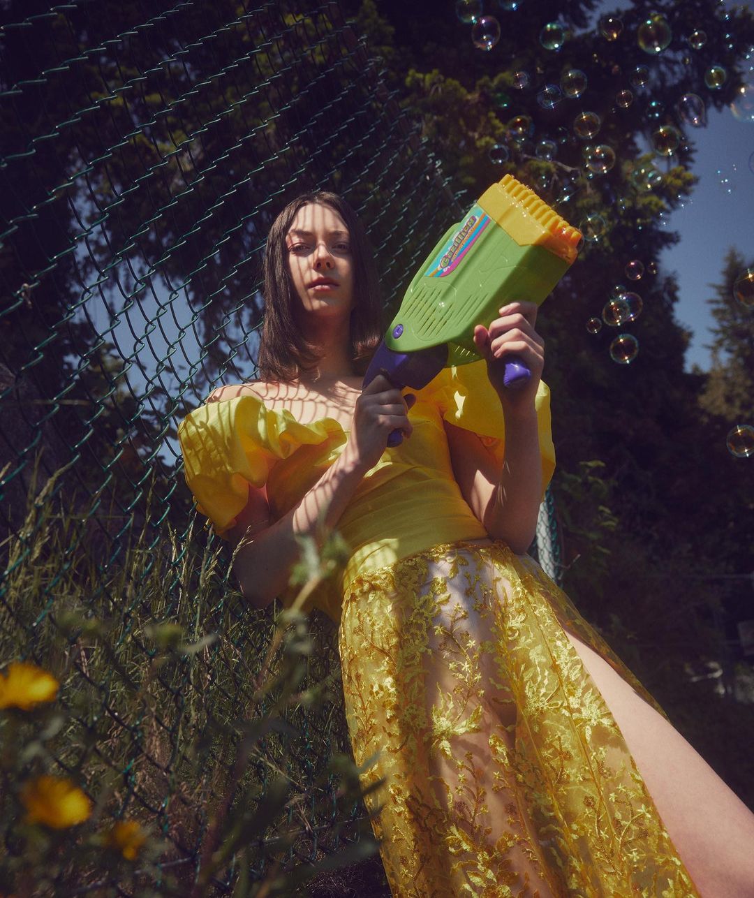 Woman in yellow lace gown holds bubble gun outdoors, fence and greenery in background, creating whimsical, playful scene with natural light.