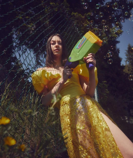 Woman in yellow lace gown holds bubble gun outdoors, fence and greenery in background, creating whimsical, playful scene with natural light.