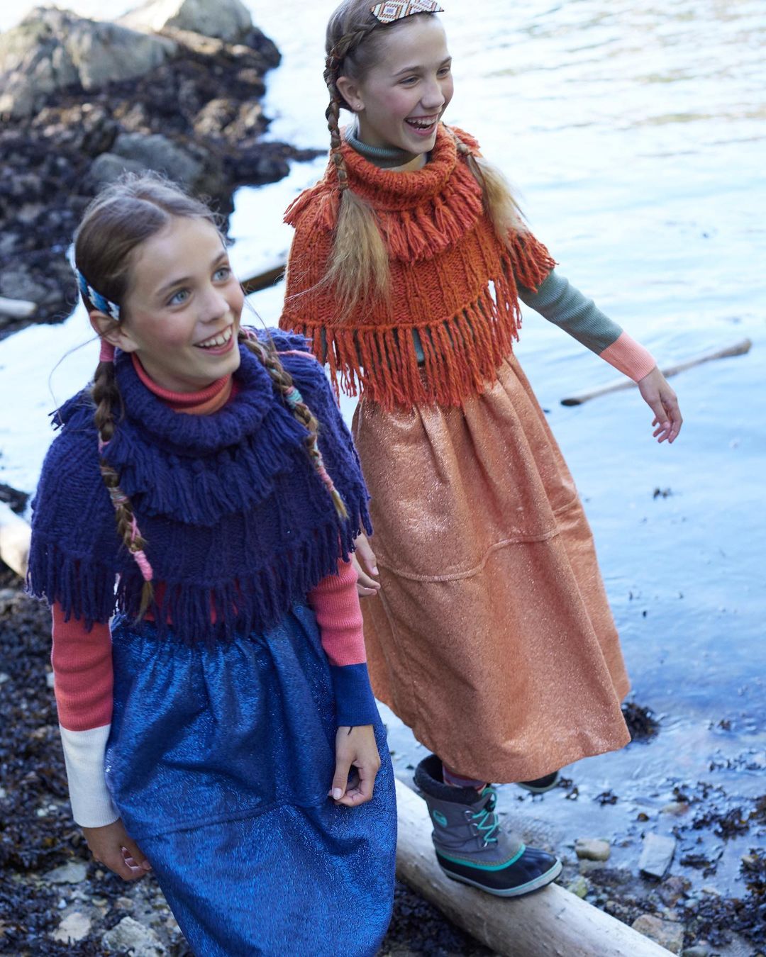 Two smiling girls wearing knitted shawls and shiny skirts, posing outdoors near a body of water with natural, soft lighting.