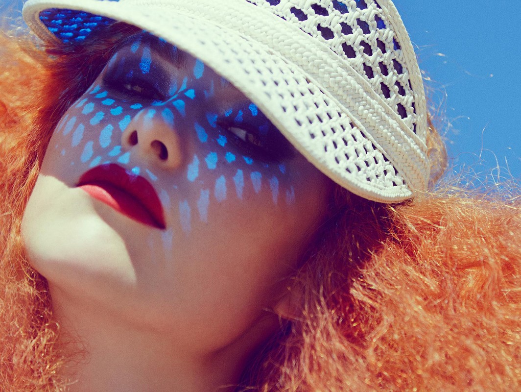 Close-up of a fashion model with red hair wearing a white visor and bold red lipstick, posing against a vibrant blue sky background.