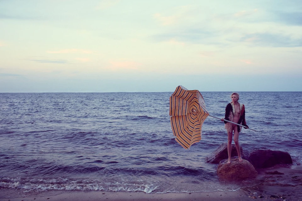 Woman standing on a rock near the ocean with a striped umbrella during sunset. Tranquil coastal scene with a fashion element.