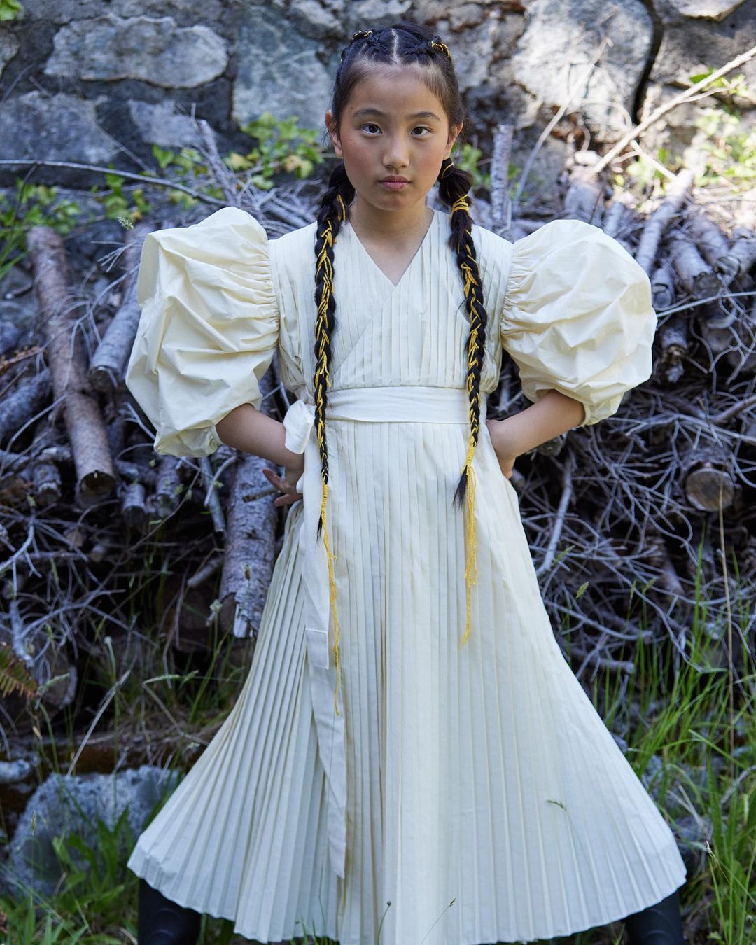 Young girl with braided hair wearing a white pleated dress with voluminous puff sleeves standing outdoors