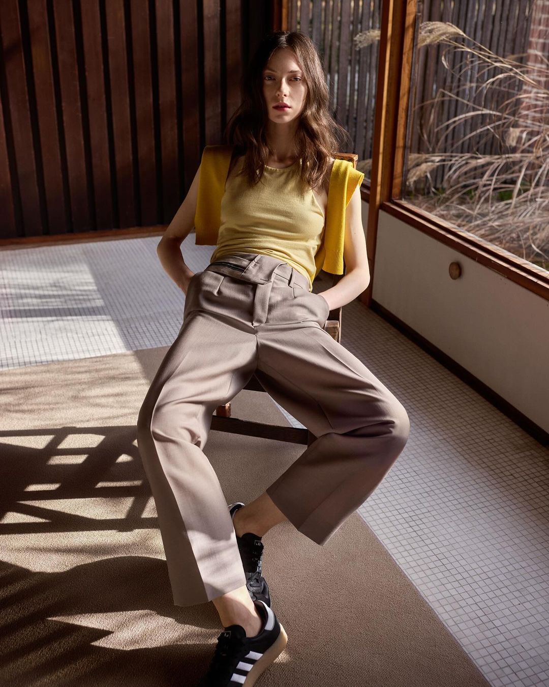 A slim young woman is sitting on a chair indoors wearing beige culottes, a light yellow top, and black sneakers in a relaxed pose.