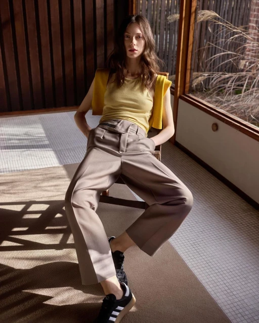 A slim young woman is sitting on a chair indoors wearing beige culottes, a light yellow top, and black sneakers in a relaxed pose.