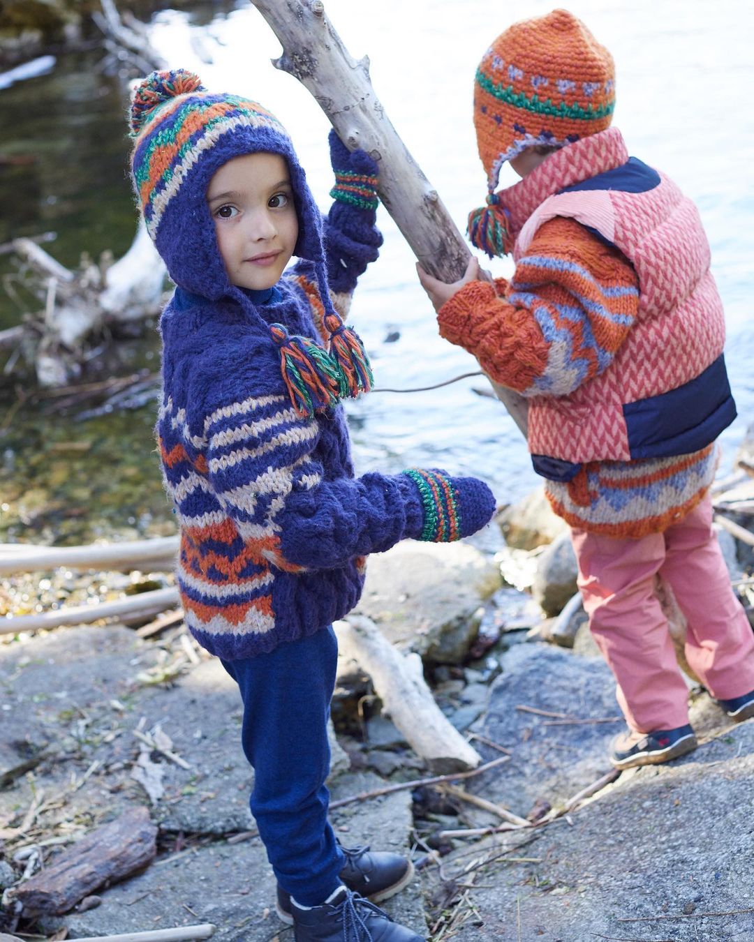 Two children in colorful knitwear stand by a tree branch near a lake, one looking at the camera, the other facing away, outdoors.