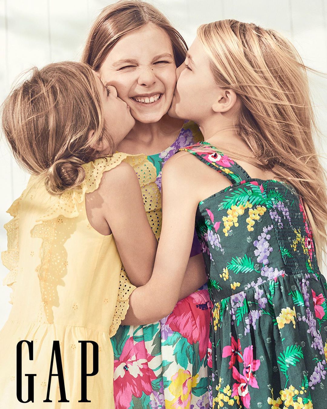 Three young girls in colorful floral dresses hug and smile; two of the girls are kissing the third girl's cheek. GAP logo is in the lower left.