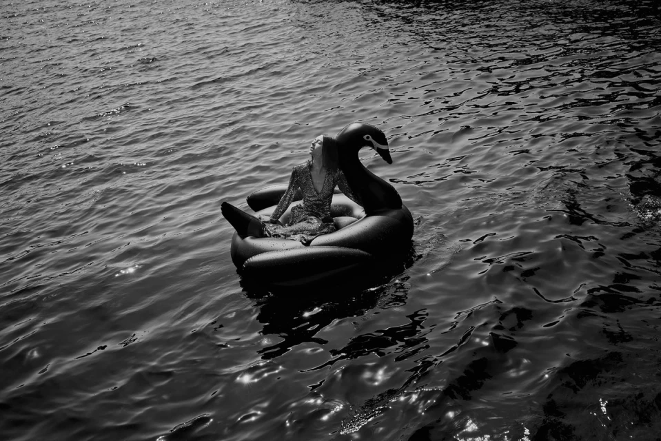 Woman in glittery dress sits atop an inflatable swan on rippling water, monochrome, outdoor setting with natural light creating reflections.