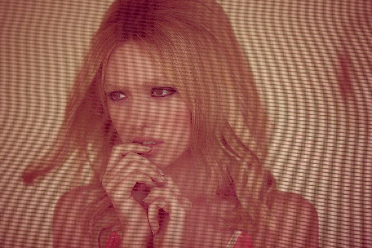Close-up portrait of a young woman with blond wavy hair, resting her hands on her chin. She has her dark eyes open, looking off to the side.