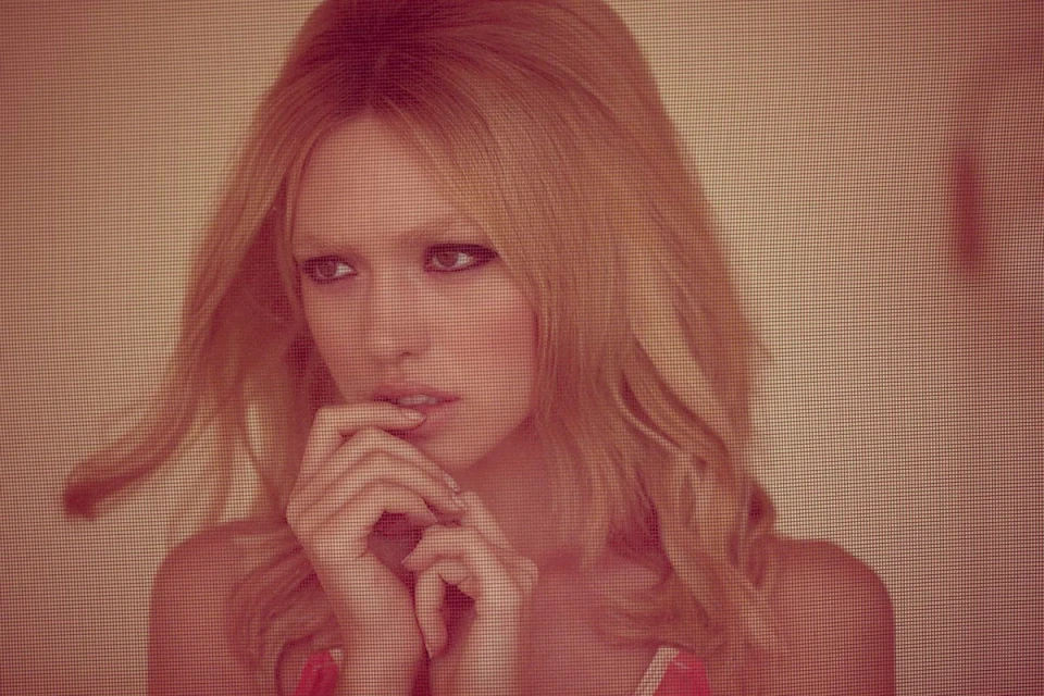 Close-up portrait of a young woman with blond wavy hair, resting her hands on her chin. She has her dark eyes open, looking off to the side.