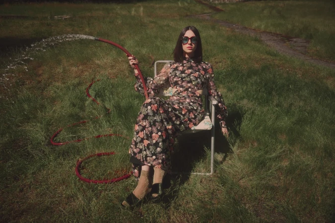 Woman in floral dress sitting in a metal chair outdoors holding a red garden hose spraying water