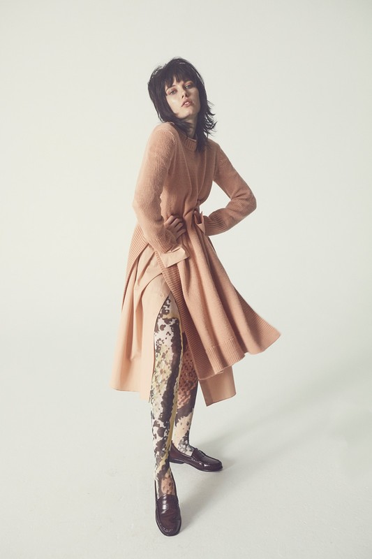 A fashionable woman poses in a light peach knit dress, patterned stockings, and shiny brown loafers against a seamless white backdrop.