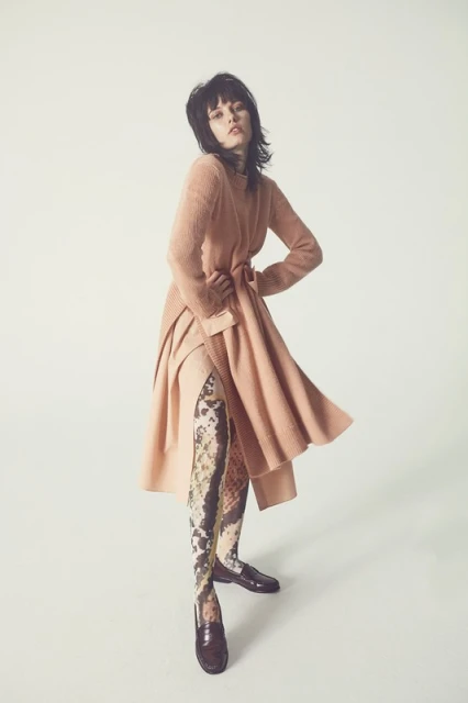 A fashionable woman poses in a light peach knit dress, patterned stockings, and shiny brown loafers against a seamless white backdrop.