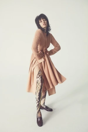 A fashionable woman poses in a light peach knit dress, patterned stockings, and shiny brown loafers against a seamless white backdrop.