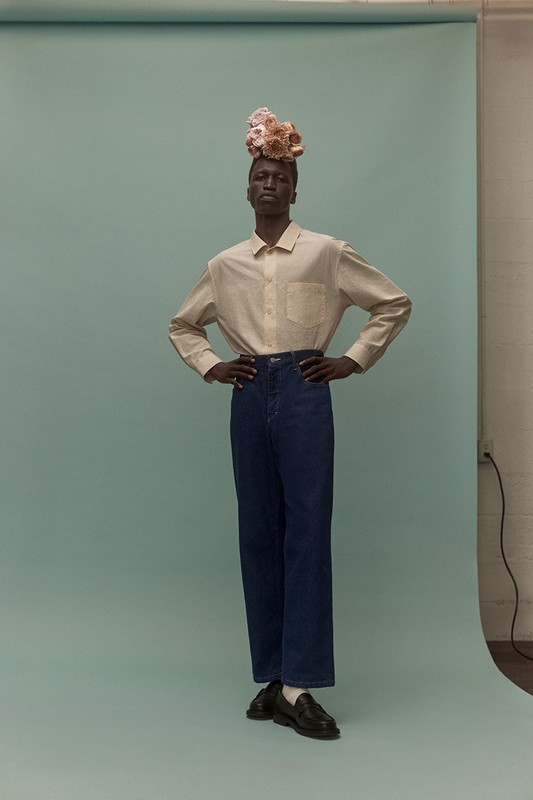 Full-body studio portrait of a Black male model wearing a button-down shirt, wide-legged jeans, loafers, and a floral headpiece.