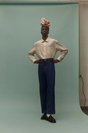 Full-body studio portrait of a Black male model wearing a button-down shirt, wide-legged jeans, loafers, and a floral headpiece.