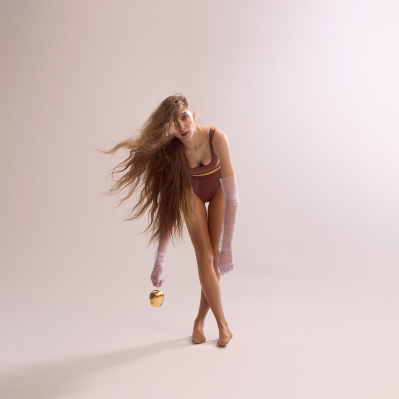 Young woman in bodysuit and sheer sleeves poses with golden apple against neutral backdrop with long flowing hair in studio setting.