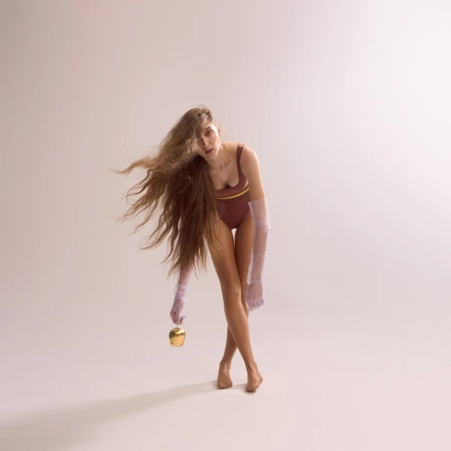 Young woman in bodysuit and sheer sleeves poses with golden apple against neutral backdrop with long flowing hair in studio setting.