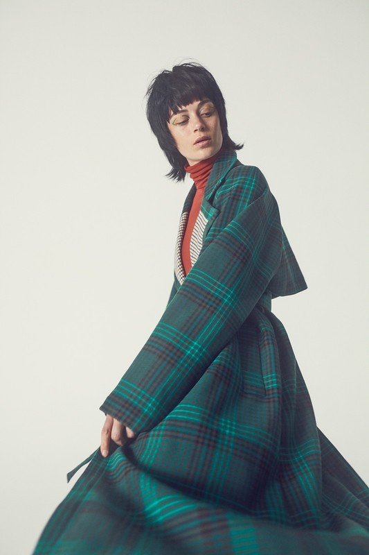 Brunette woman with blunt bangs poses wearing a green and dark red plaid coat on a white background in a fashion setting.