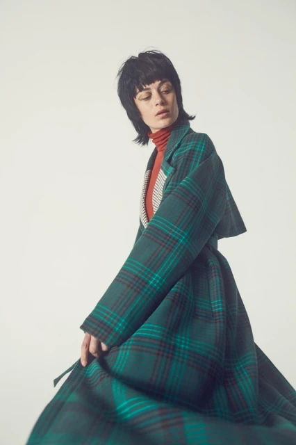 Brunette woman with blunt bangs poses wearing a green and dark red plaid coat on a white background in a fashion setting.