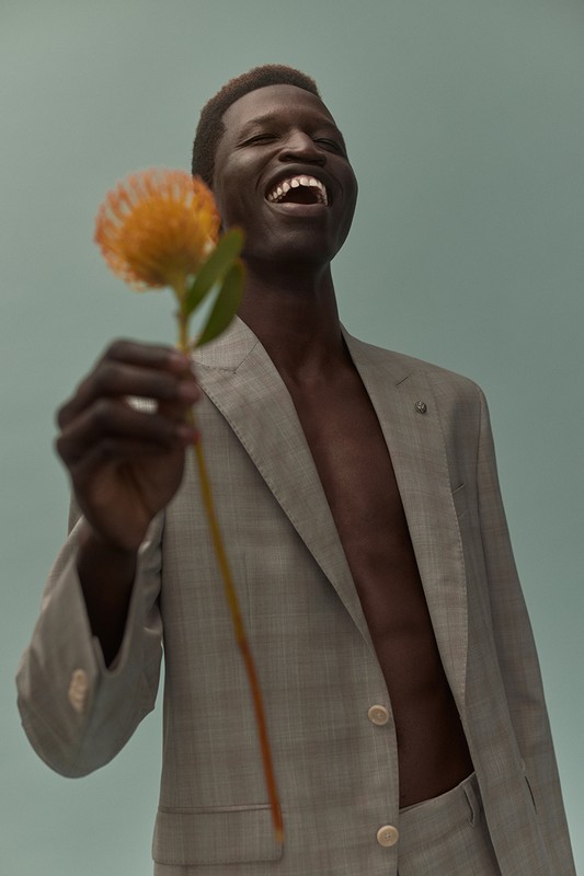 Smiling Black man in a grey suit holds out a peach-colored pincushion flower against a light blue background in a fashion portrait.