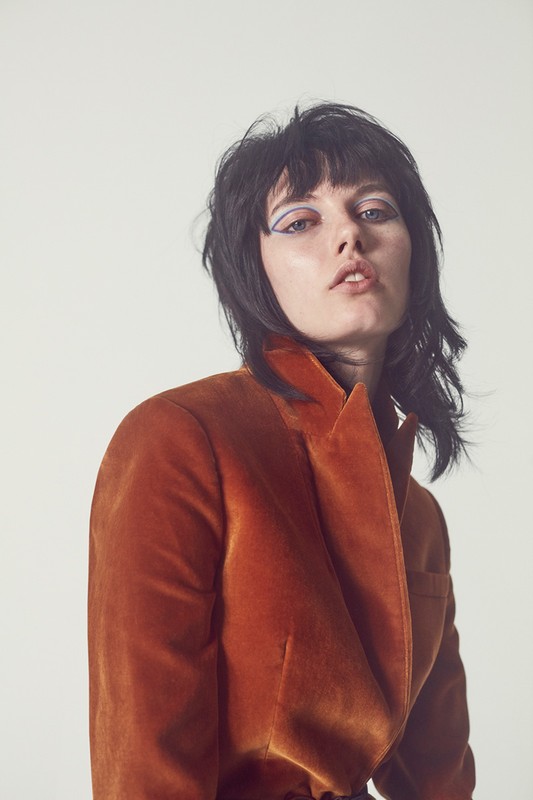 Portrait of a female model in a rust-colored velvet jacket with blue eyeshadow, shot against a white backdrop in a studio setting.