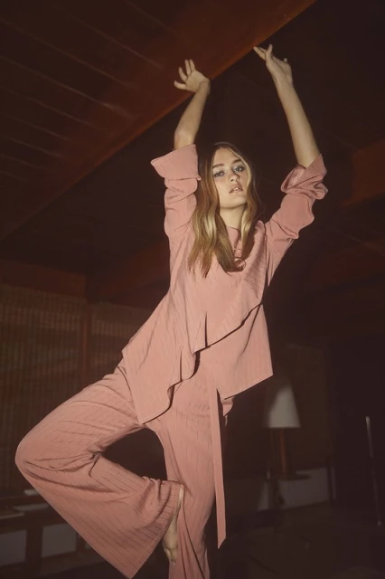 Woman in pink pantsuit strikes a dynamic pose indoors, illuminated by a lamp. The setting is characterized by a vintage aesthetic.