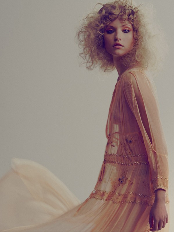 Portrait of an adult female fashion model with curly blond hair wearing a flowy, peach-colored dress in a studio setting with soft lighting.