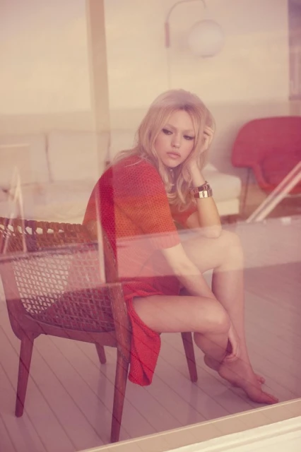 Young woman in a red dress sits barefoot on a wicker chair by the window, bathed in soft light, exuding a relaxed and contemplative mood.