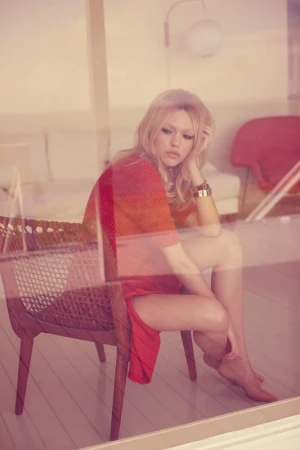 Young woman in a red dress sits barefoot on a wicker chair by the window, bathed in soft light, exuding a relaxed and contemplative mood.