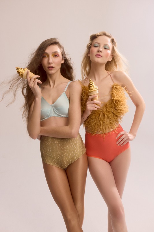 Two women pose in a studio wearing lingerie and holding golden ice cream cones. The shot is brightly lit with a soft, dreamy quality.