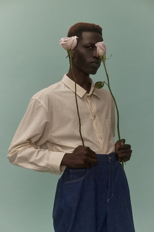 Portrait of dark-skinned male model holding pink roses against light blue background in studio. Man wears collared shirt and blue denim pants.