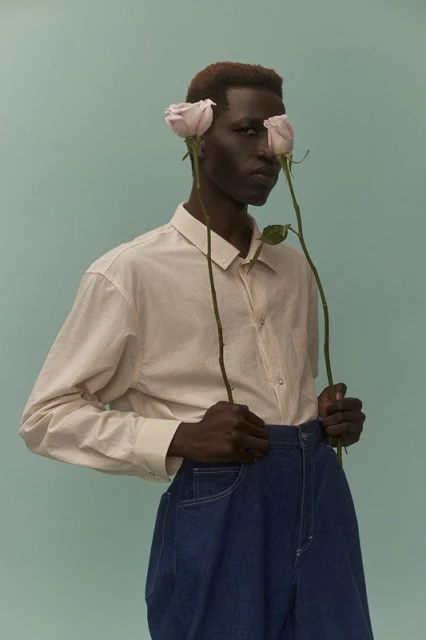 Portrait of dark-skinned male model holding pink roses against light blue background in studio. Man wears collared shirt and blue denim pants.