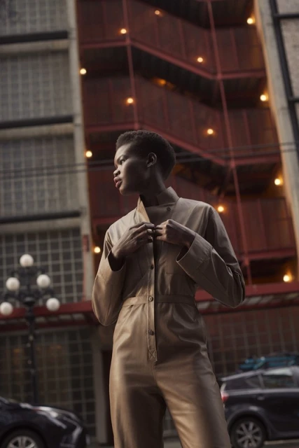 African woman in a beige jumpsuit on a city street, looking left with car and building in background, with orange lighting.