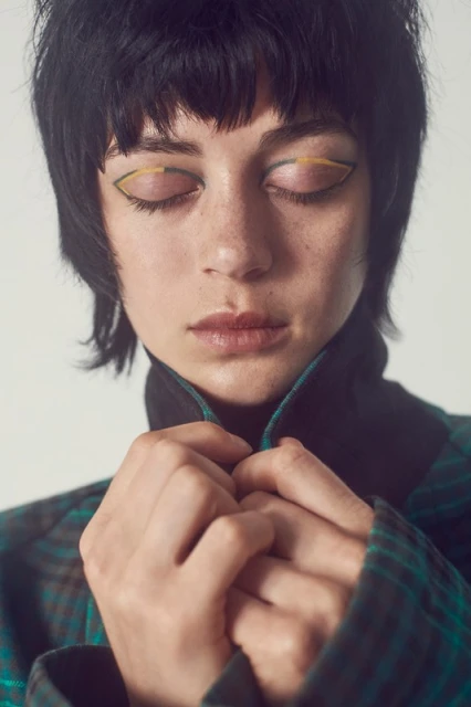 Close-up of a young woman with dark hair, wearing a plaid top with a black collar, and yellow and green eyeshadow with closed eyes.