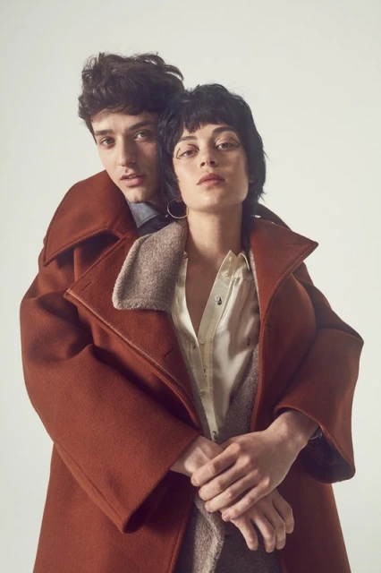 A young couple embraces in a studio shot, wearing a rust-colored wool coat and shirt, with a plain off-white background creating soft, diffused lighting.
