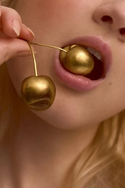 Close-up of an adult woman with blonde hair holding golden decorative cherries to her mouth, beauty, fashion, and food-related content.