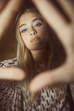 Close-up portrait of a young woman framed by out-of-focus hands, wearing blue eyeshadow and a printed top.