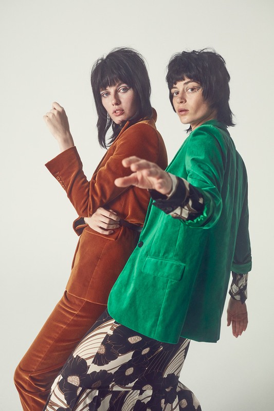 Two fashion models pose indoors against a white background wearing a copper velvet suit and a green velvet jacket and floral skirt.