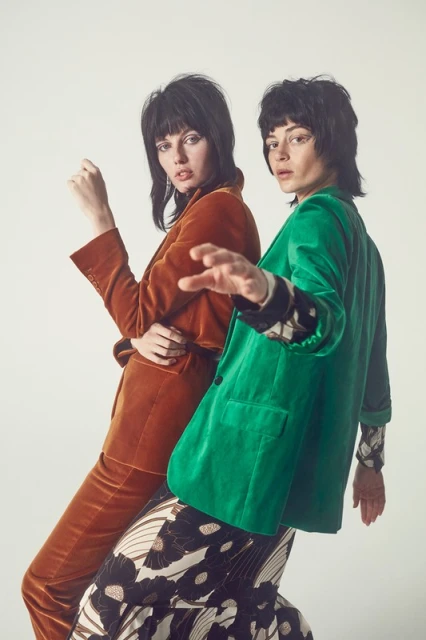 Two fashion models pose indoors against a white background wearing a copper velvet suit and a green velvet jacket and floral skirt.
