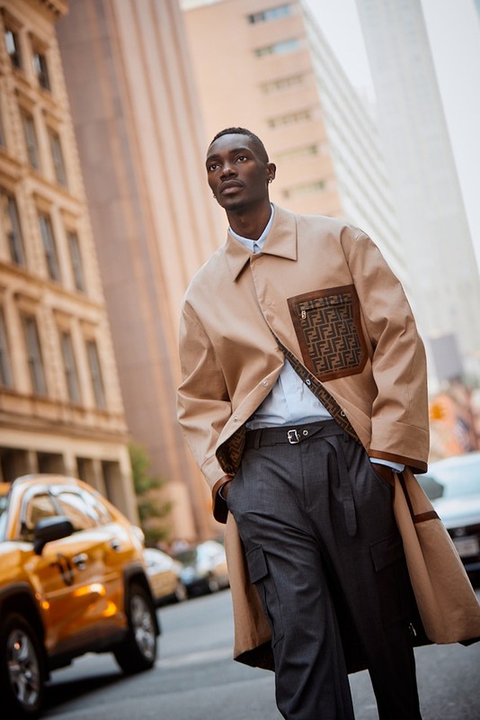 An adult man walks on an urban street, wearing a tan coat and grey trousers. He is walking past a yellow taxi cab.