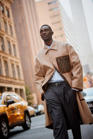 An adult man walks on an urban street, wearing a tan coat and grey trousers. He is walking past a yellow taxi cab.