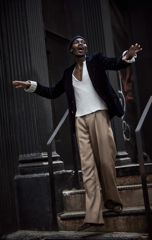 Stylish African American man stands on outdoor steps, wearing a blazer, trousers, beanie, and white v-neck, with his arms outstretched.