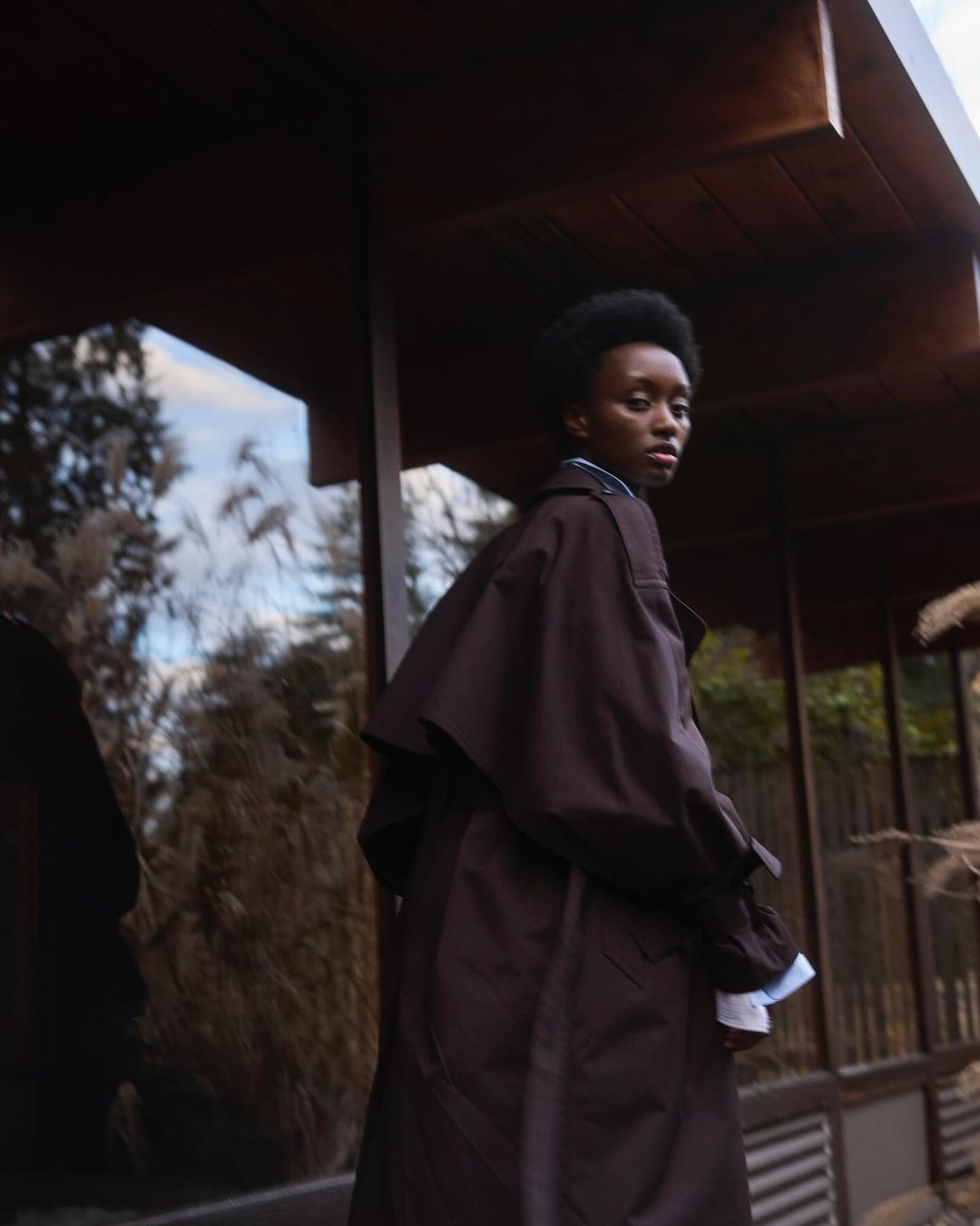 An African-American woman in a brown trench coat stands outside a modern building, glancing behind her with a serene expression.