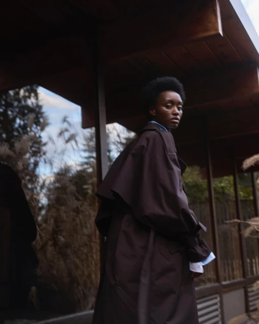 An African-American woman in a brown trench coat stands outside a modern building, glancing behind her with a serene expression.